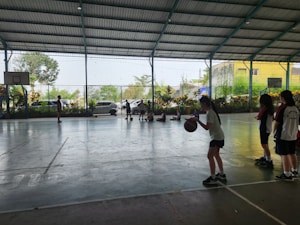 A group of young people are inside an indoor sports court. Several children are engaged in activity, with some playing basketball and others sitting on the floor against the fence. Greenery is visible outside through the netting.