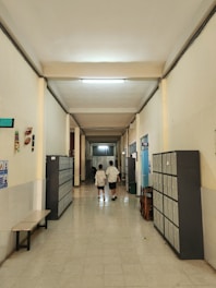 A school corridor with tiled floors and high ceilings, lined with lockers on both sides. Two individuals wearing similar patterned clothing walk away from the camera down the hallway. There are benches and chairs along the sides, and some posters and decorations on the walls.