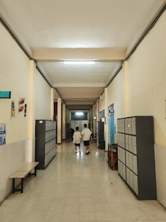 A school corridor with tiled floors and high ceilings, lined with lockers on both sides. Two individuals wearing similar patterned clothing walk away from the camera down the hallway. There are benches and chairs along the sides, and some posters and decorations on the walls.