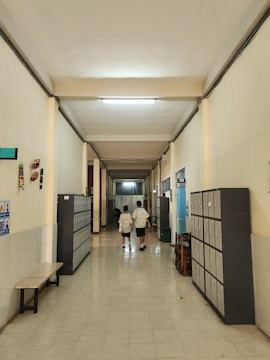 A school corridor with tiled floors and high ceilings, lined with lockers on both sides. Two individuals wearing similar patterned clothing walk away from the camera down the hallway. There are benches and chairs along the sides, and some posters and decorations on the walls.