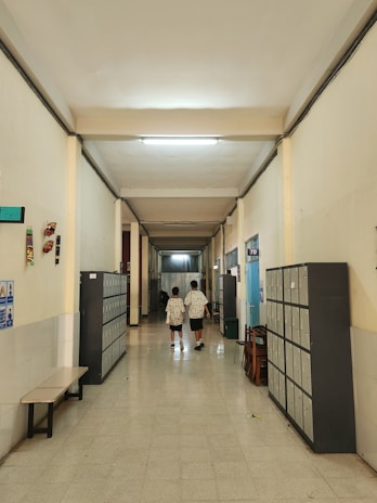 A school corridor with tiled floors and high ceilings, lined with lockers on both sides. Two individuals wearing similar patterned clothing walk away from the camera down the hallway. There are benches and chairs along the sides, and some posters and decorations on the walls.