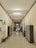 A school corridor with tiled floors and high ceilings, lined with lockers on both sides. Two individuals wearing similar patterned clothing walk away from the camera down the hallway. There are benches and chairs along the sides, and some posters and decorations on the walls.