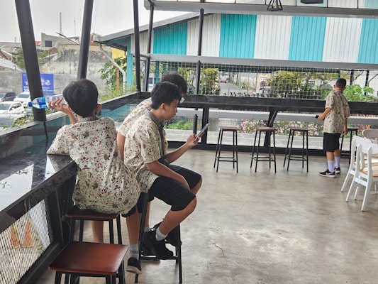 A group of people wearing matching floral shirts sits and stands in a casual, open-air setting. One person drinks from a bottle, another looks at a phone, while another stands near the railing. Various stools and a metal mesh barrier are part of the modern, industrial decor. Large windows open to a view of a building with turquoise and white stripes, and there are plants and parked cars visible outside.