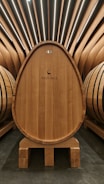 A large wooden barrel with horizontal black bands sits prominently in the center of a room, surrounded by similar barrels. The wood has a rich, warm tone, and the top of the barrel is marked with 'Arzuaga' and a deer logo. The room features a modern design with sleek, angled wooden slats that converge at the ceiling, creating an elegant and sophisticated atmosphere.
