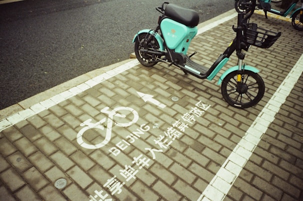 A teal electric scooter is parked on a designated bike parking area marked with white paint symbols and text in both English and Chinese. The ground consists of paved bricks, and the area appears to be part of a street sidewalk.
