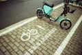 A teal electric scooter is parked on a designated bike parking area marked with white paint symbols and text in both English and Chinese. The ground consists of paved bricks, and the area appears to be part of a street sidewalk.
