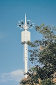 A tall, white telecommunications tower stands against a clear blue sky. The tower's structure features multiple antennas at the top. Green foliage is partially visible, hinting at a natural setting.