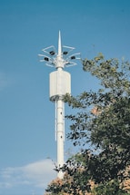A tall, white telecommunications tower stands against a clear blue sky. The tower's structure features multiple antennas at the top. Green foliage is partially visible, hinting at a natural setting.