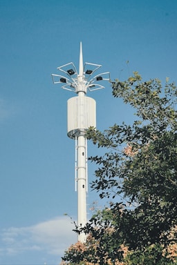 A tall, white telecommunications tower stands against a clear blue sky. The tower's structure features multiple antennas at the top. Green foliage is partially visible, hinting at a natural setting.
