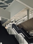 A modern interior with a geometric ceiling design featuring exposed beams and panels. A wide staircase with illuminated railings leads upwards, and a concrete pillar supports the structure. The area has a minimalist aesthetic with clean lines and an industrial feel.