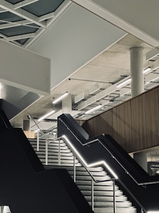 A modern interior with a geometric ceiling design featuring exposed beams and panels. A wide staircase with illuminated railings leads upwards, and a concrete pillar supports the structure. The area has a minimalist aesthetic with clean lines and an industrial feel.