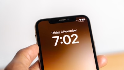 A hand holding a smartphone that displays the date as Friday, 3 November and the time as 7:02. The screen has a simple design with white text on a gradient brown background.