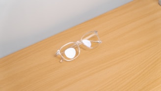 A pair of transparent eyeglasses with clear lenses is placed on a flat, light brown wooden surface. The eyeglasses are positioned at an angle, with the earpieces slightly spread apart.