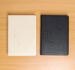 Two notebooks are placed on a wooden surface. The one on the left is light beige with a subtle dandelion pattern and an embossed illustration. The one on the right is black with a similar dandelion design.