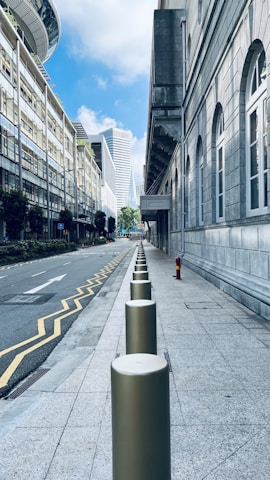 A city street scene featuring a row of bollards aligned along a wide stone sidewalk. The street is bordered by modern office buildings with large windows on one side and a classic stone facade building with arched windows on the other. The sky is partly cloudy with a bright blue hue, and several skyscrapers can be seen in the background, adding a sense of urban depth.