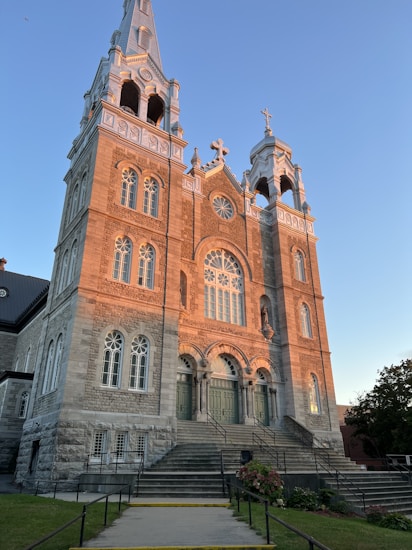 A large, ornate stone church building with two symmetrical towers featuring arched windows and decorative details, bathed in warm sunlight. The entrance is accessible by wide stone steps leading up to three large, intricately designed wooden doors. The surrounding area includes a well-maintained lawn and some blooming flowers.