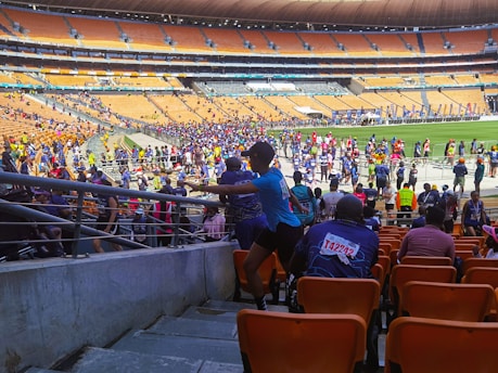 A bustling stadium filled with participants, some wearing race bibs with colorful numbers, moving along a designated path. The crowd, dressed in various athletic outfits, extends across the field and stands. The seating is mostly empty, consisting of orange seats, with a few onlookers scattered about.