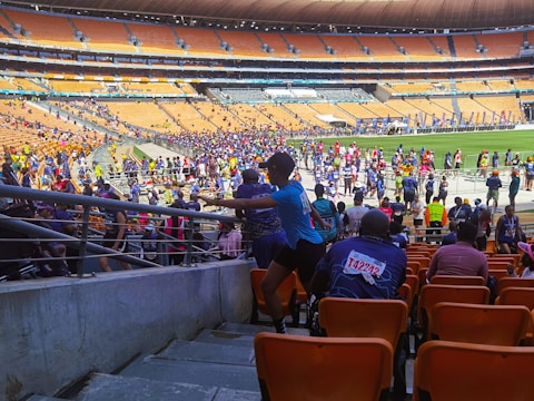 A bustling stadium filled with participants, some wearing race bibs with colorful numbers, moving along a designated path. The crowd, dressed in various athletic outfits, extends across the field and stands. The seating is mostly empty, consisting of orange seats, with a few onlookers scattered about.