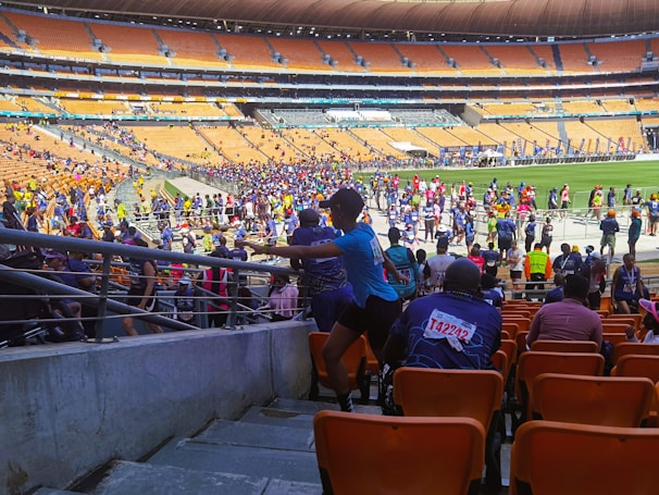 A bustling stadium filled with participants, some wearing race bibs with colorful numbers, moving along a designated path. The crowd, dressed in various athletic outfits, extends across the field and stands. The seating is mostly empty, consisting of orange seats, with a few onlookers scattered about.