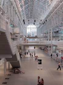 A spacious, modern indoor hall with high ceilings and large windows letting in natural light. The interior features metal staircases and walkways. People are walking and sitting in various areas, giving a sense of activity and casual sociability.
