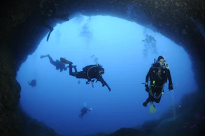 Scuba divers exploring a dramatic underwater cave illuminated by beams of sunlight.