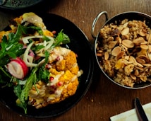 A black plate holds a vibrant dish featuring roasted cauliflower garnished with fresh greens, radish slices, pomegranate seeds, and what appears to be a creamy sauce. To the side, a metal bowl contains a rice dish mixed with sliced almonds and possibly dried fruits.