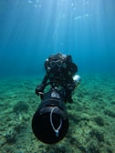 A scuba diver is underwater, navigating with a propulsion device over a rocky seabed. Sunlight pierces through the clear blue water from above, creating rays that illuminate the scene and add a sense of depth.