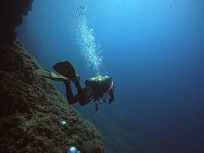 A scuba diver swimming underwater near a rocky seabed, with air bubbles rising to the surface. The scene is illuminated by the natural light filtering through the blue water, giving it an ambient and tranquil atmosphere.