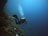 A scuba diver swimming underwater near a rocky seabed, with air bubbles rising to the surface. The scene is illuminated by the natural light filtering through the blue water, giving it an ambient and tranquil atmosphere.