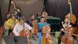 A group of people are seated and engaged in playing string instruments, particularly cellos, in a room. The setting appears to be a music class or rehearsal. There is a variety of ages among the participants, and one person wears a mask. The backdrop features a large stringed instrument, possibly a harp, and a table covered with a green cloth is visible towards the back.