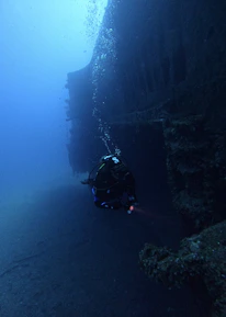 A diver is exploring an underwater shipwreck, surrounded by deep blue water. The wreck is encrusted with marine growth, and bubbles from the diver's equipment are rising to the surface.