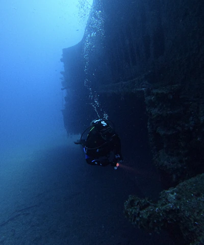 A diver is exploring an underwater shipwreck, surrounded by deep blue water. The wreck is encrusted with marine growth, and bubbles from the diver's equipment are rising to the surface.