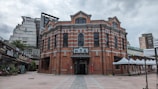 A historic red brick building with elegant architecture featuring arched windows and decorative patterns. It stands prominently in an open plaza surrounded by modern buildings. White tents line the right side of the plaza, and signage is visible indicating commercial activity in the area.