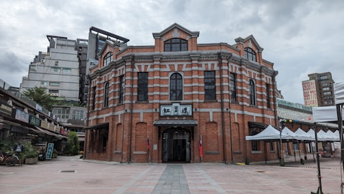 A historic red brick building with elegant architecture featuring arched windows and decorative patterns. It stands prominently in an open plaza surrounded by modern buildings. White tents line the right side of the plaza, and signage is visible indicating commercial activity in the area.