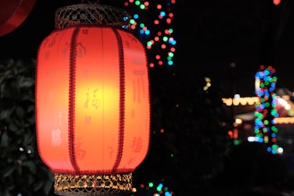 A traditional red lantern with Chinese characters is illuminated against a dark background. Bright, colorful lights are blurred in the distance, creating a festive atmosphere.