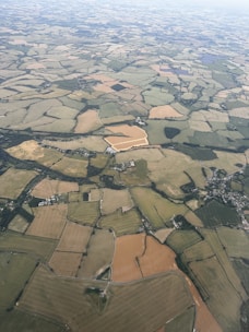 Aerial view of vast, patchwork farmland showing a variety of fields in different stages of cultivation, interspersed with clusters of trees and small settlements. The landscape is characterized by irregularly shaped plots of land, with variations in color indicating different types of crops or grasslands.