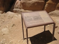 A sandy, rocky landscape features an informational sign on a brown metal stand, displaying text and images related to the surroundings. The background consists of large, smooth rock formations with varying shades of beige and brown.