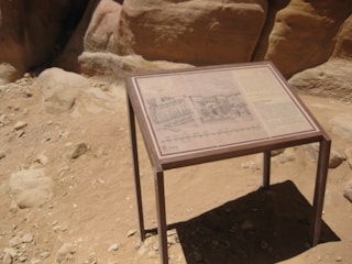 A sandy, rocky landscape features an informational sign on a brown metal stand, displaying text and images related to the surroundings. The background consists of large, smooth rock formations with varying shades of beige and brown.