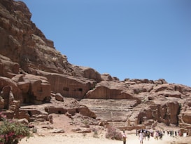 A large rocky landscape with ancient carved structures resembling an amphitheater or tombs. The sand-colored rock formations rise steeply against a clear blue sky. Groups of tourists are seen at the base, indicating a popular historical or archaeological site. Sparse vegetation with small bushes or flowers is visible in the foreground.
