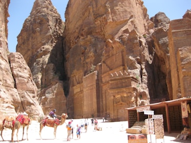 A group of camels with colorful saddle decorations are guided by people near a large, intricately carved rock facade in a desert setting. The façade is part of steep, towering cliffs under a bright blue sky. A small market stall with signs is set up nearby, selling goods to visitors.