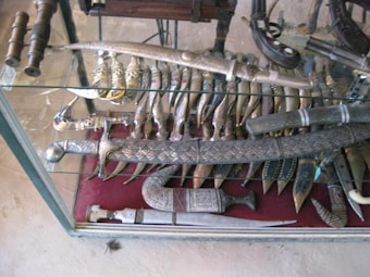 A display case containing an assortment of traditional knives and swords. The knives appear to be ornate with detailed handles and some with curved blades. The swords have intricate designs on their hilts and scabbards. The weapons are arranged neatly on a red velvet surface within the glass case.