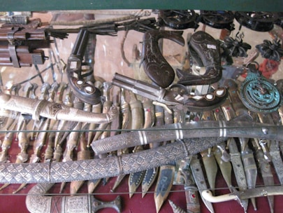 A collection of antique weapons and ornate daggers displayed inside a glass case. The items include intricately designed knives, swords, and what appear to be vintage firearms with decorative handles and inlays.