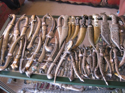 An assortment of intricately designed daggers with curved, ornate handles and scabbards displayed on a glass surface. The daggers vary in size and feature detailed engravings and patterns, showcasing craftsmanship in metalwork.