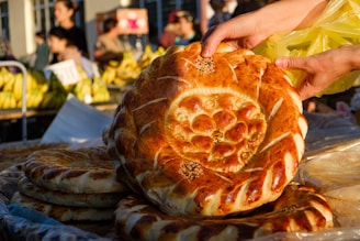 A person is holding a large, round, golden-brown bread with decorative patterns on top. More loaves of similar bread are stacked underneath it. In the background, there are bananas for sale, and several people are mingling in a market setting.
