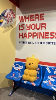 A yellow teddy bear with a small pointed hat sits on a row of blue plastic seats adorned with multiple 'BUTTER' stickers. A wall behind displays the large text 'WHERE IS YOUR HAPPINESS' in red, followed by 'BETTER LIFE, BETTER BUTTER' in blue. A decorative round sign is affixed to the adjacent yellow wall.