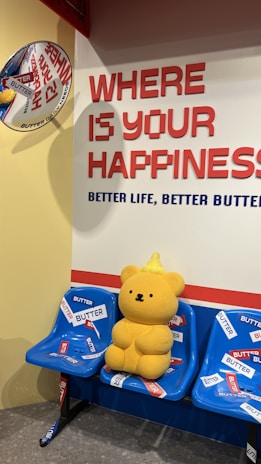 A yellow teddy bear with a small pointed hat sits on a row of blue plastic seats adorned with multiple 'BUTTER' stickers. A wall behind displays the large text 'WHERE IS YOUR HAPPINESS' in red, followed by 'BETTER LIFE, BETTER BUTTER' in blue. A decorative round sign is affixed to the adjacent yellow wall.