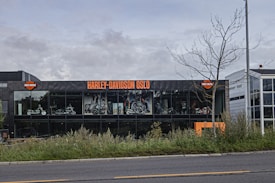 A modern Harley-Davidson dealership in Oslo with large glass windows displays motorcycles inside. The building has a sleek black facade with bright orange signage. The foreground features grass and a partially bare tree, set against a cloudy sky.