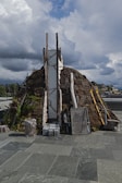 A rustic, dome-shaped structure made of stones, wood, and plant life sits on a paved surface. The sky above is filled with dramatic clouds, suggesting the approach of a storm or turbulent weather. Various items including logs, ladders, and panels are leaned against the structure. Nearby structures and trees can be seen in the background.
