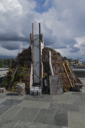 A rustic, dome-shaped structure made of stones, wood, and plant life sits on a paved surface. The sky above is filled with dramatic clouds, suggesting the approach of a storm or turbulent weather. Various items including logs, ladders, and panels are leaned against the structure. Nearby structures and trees can be seen in the background.