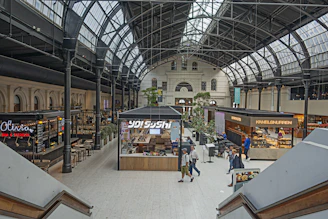 A spacious indoor marketplace with high, arched ceilings and large windows providing ample natural light. The scene includes various food stalls with visible signage, seating areas, and people walking or standing. The architecture has an industrial and historic style, combining metal beams and glass. Green plants add an element of nature to the space.